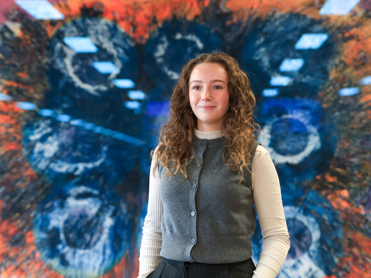 A woman with mid length curly brown hair dressed in a grey vest.