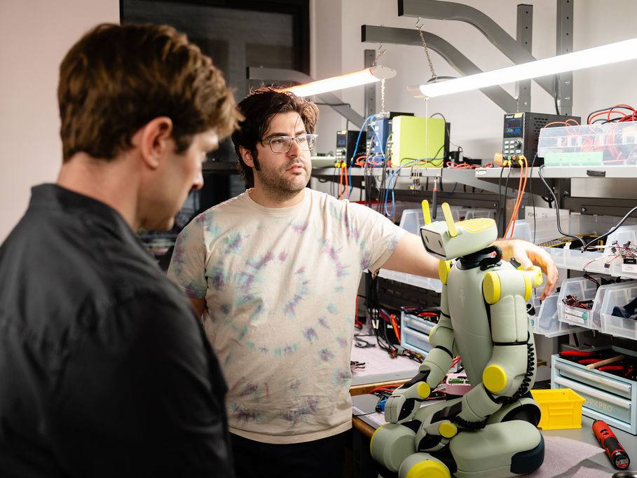 Two men standing next to a short humanoid robot sitting on a workbench with overhead lights.