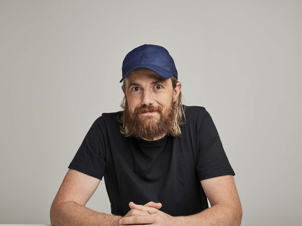 A middle-aged man with long hair and a beard, wearing a t shirt and baseball cap, poses at a desk for the camera.