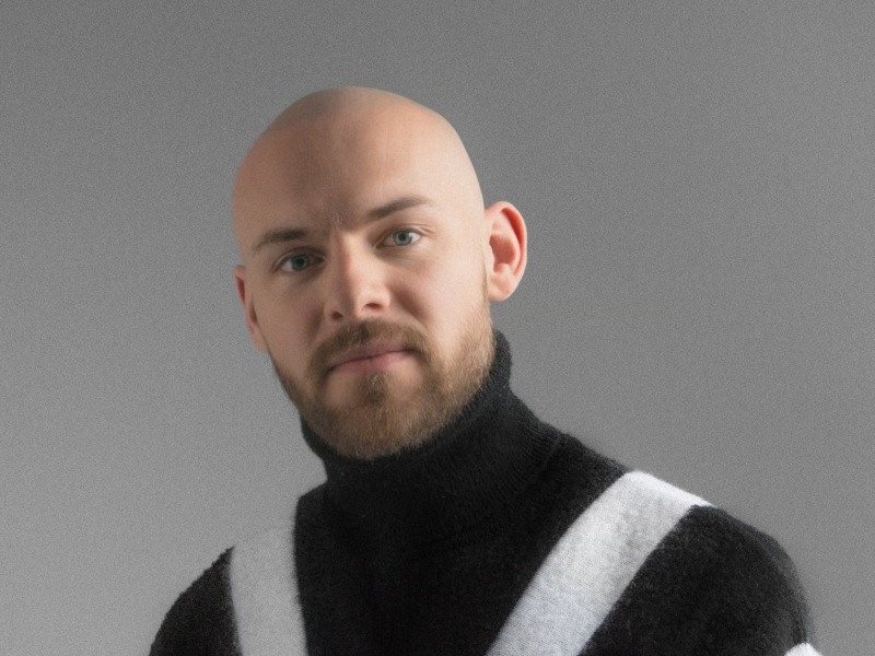 A headshot of Jeremy Bradley. He wears a high-necked wool jumper of black with a white stripe and looks seriously at the camera, against a grey background.