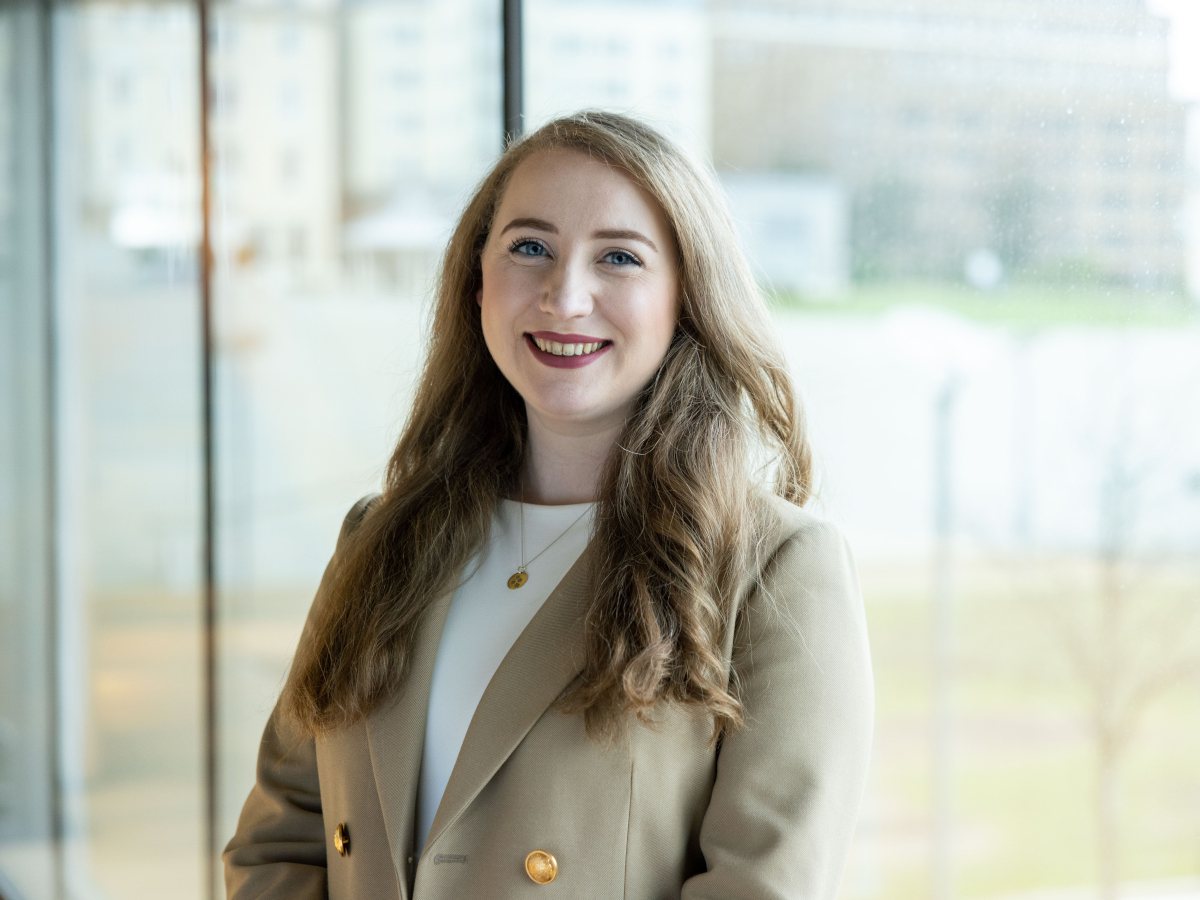 A woman wearing a tan suit jacket over a white top smiles at the camera in front of a window.