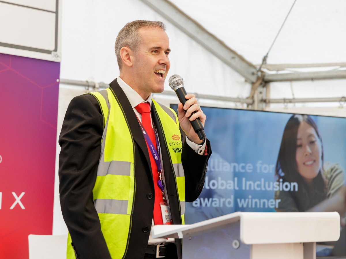 A man in a suit has a high-vis jacket on top of it. He is speaking into a mic.