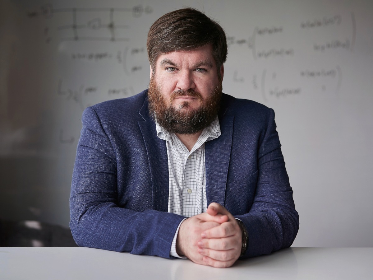 A man in a blue blazer and white shirt sitting with his arms folded on a table in front of him.