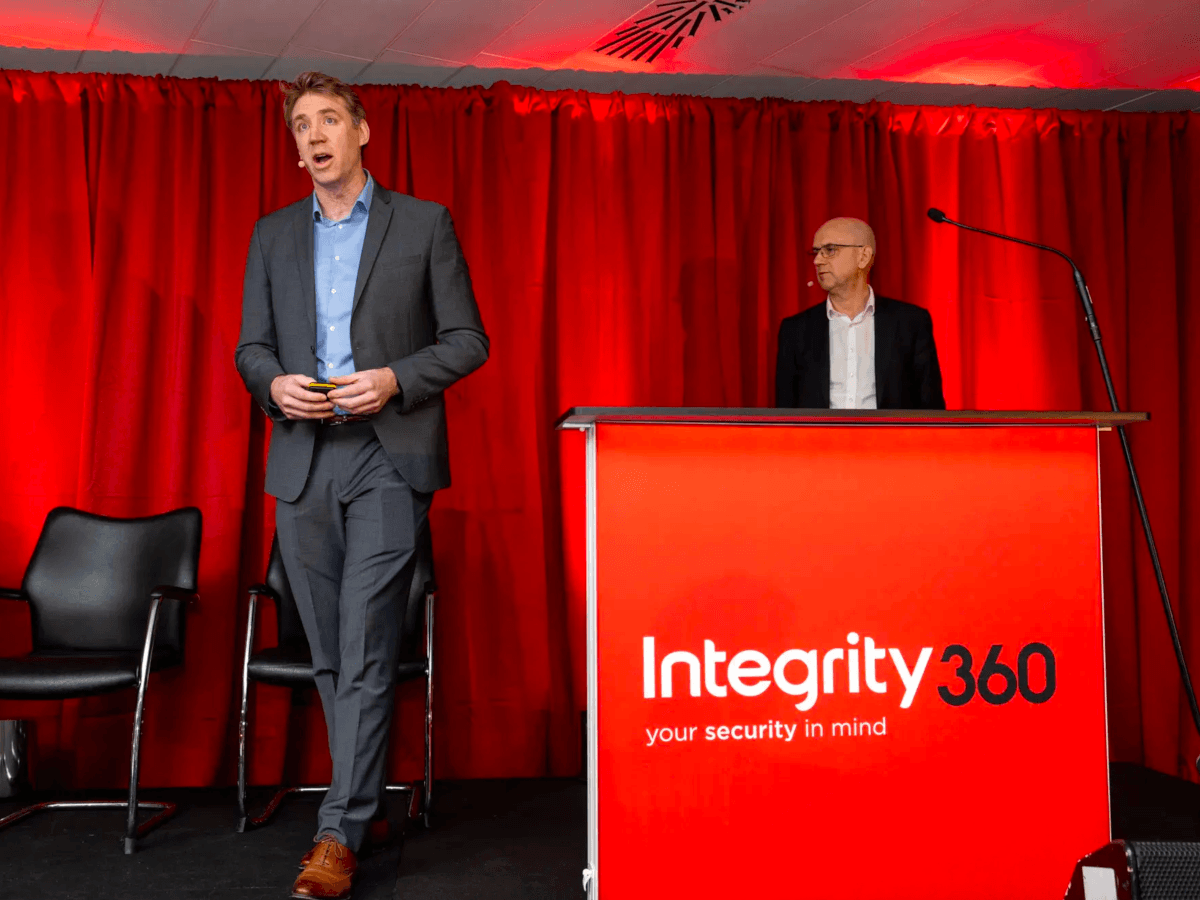Two men wearing suits stand on a stage with a red curtain and a red podium displaying the Integrity360 logo.