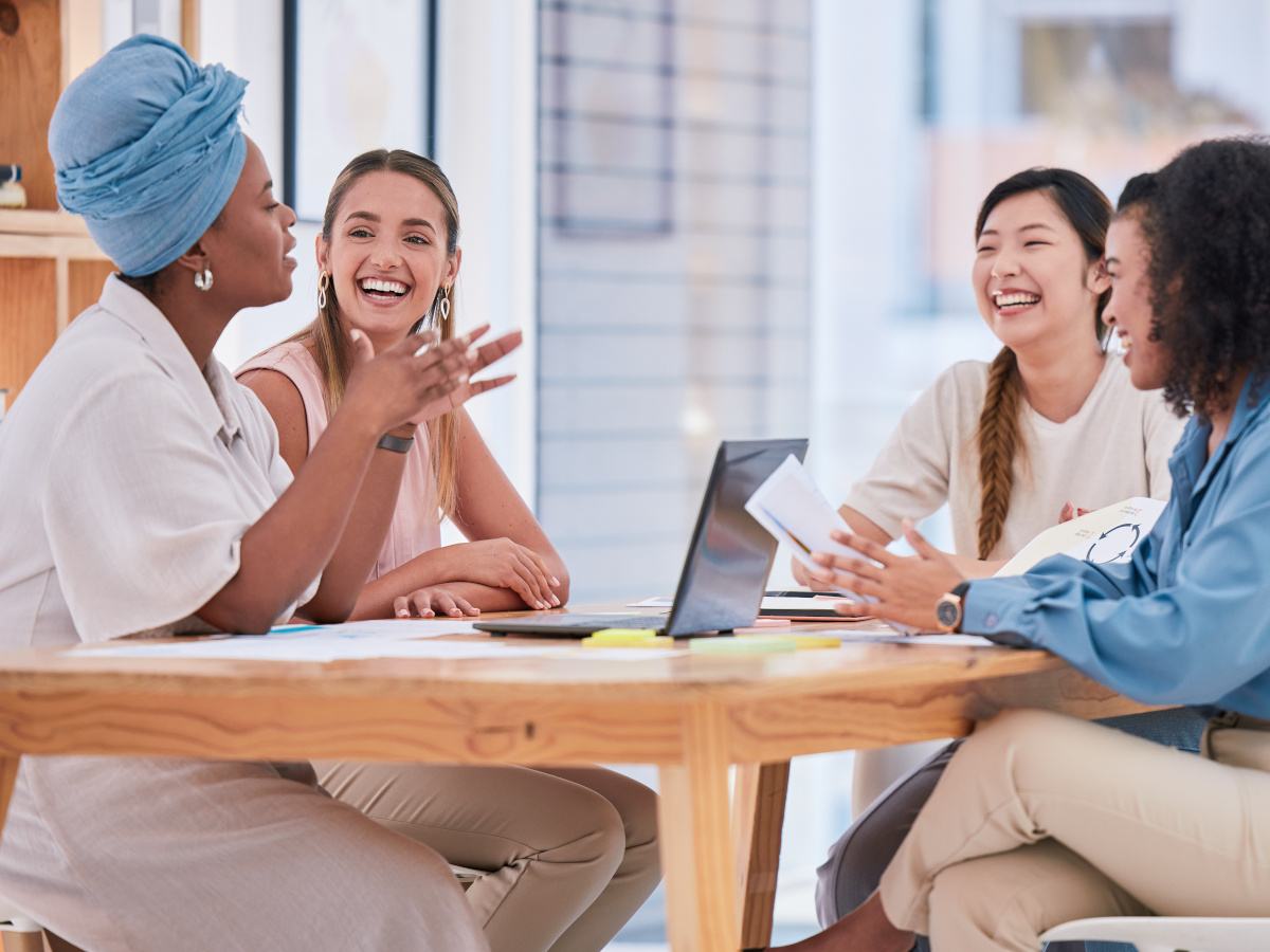 Four racially diverse young women sit around a table and talk.