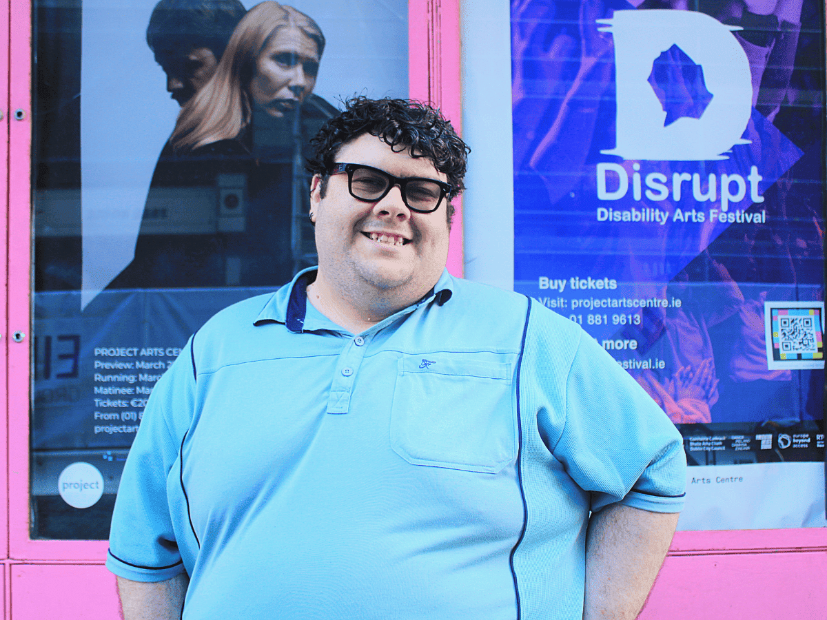 A man in a blue T-shirt smiling at the camera. He is wearing black-rimmed glasses and has curly black hair.
