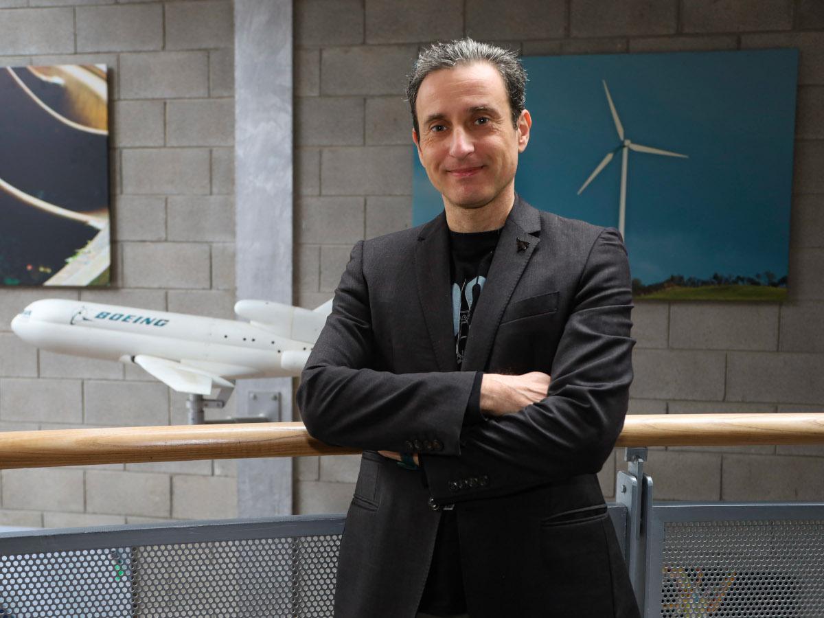 A man wearing a black suit jacket over a dark t-shirt smiles with his arms folded while standing in front of a model of an aircraft.