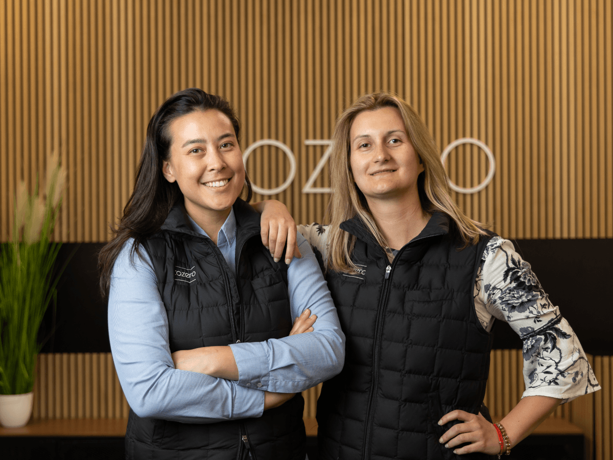 A dark-haired woman and blonde woman pose in front of the logo of their company, Tozero.