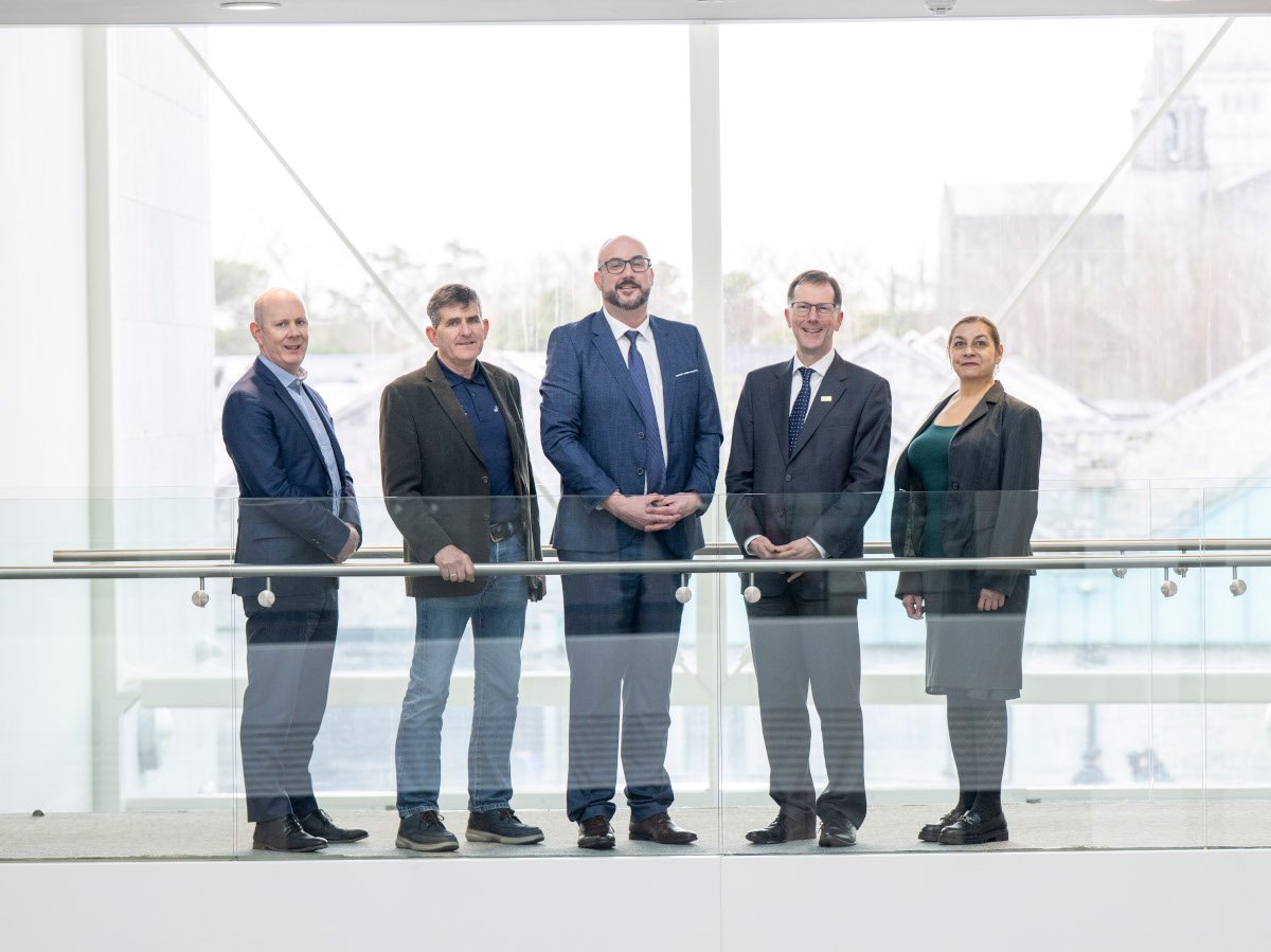 Five people in suits standing afar in what looks to be a corridor with large windows.