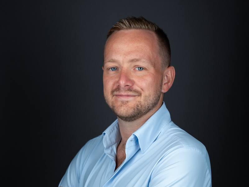 A man wearing a blue shirt with the collar open smiles at the camera in front of a dark background.
