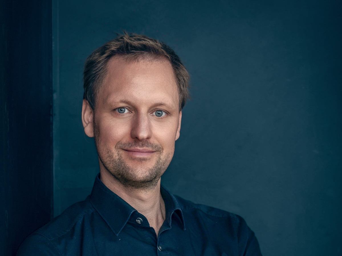 A young, white, blond man in a navy shirt posing against a teal wall.