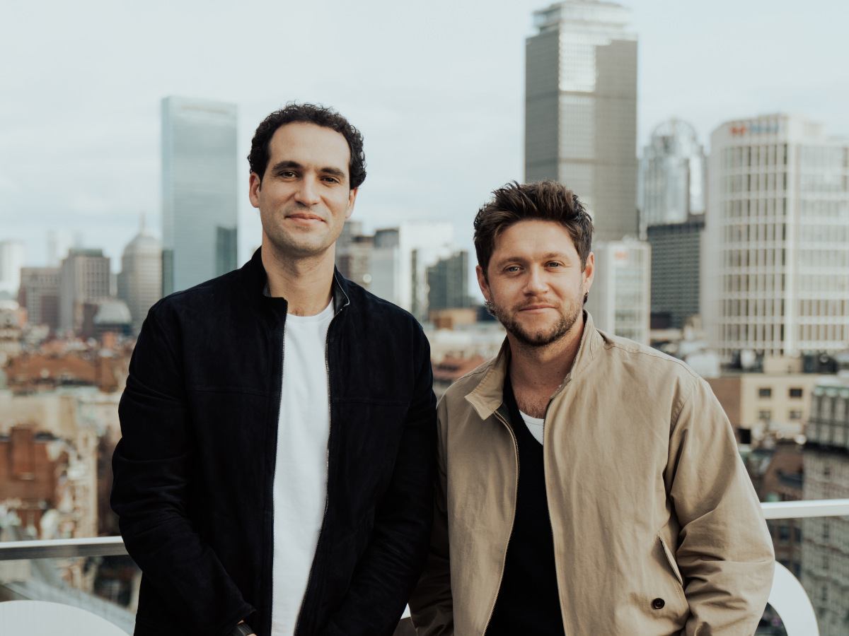 Two men wearing casual clothes stand together smiling in front of a balcony overlooking a cityscape.
