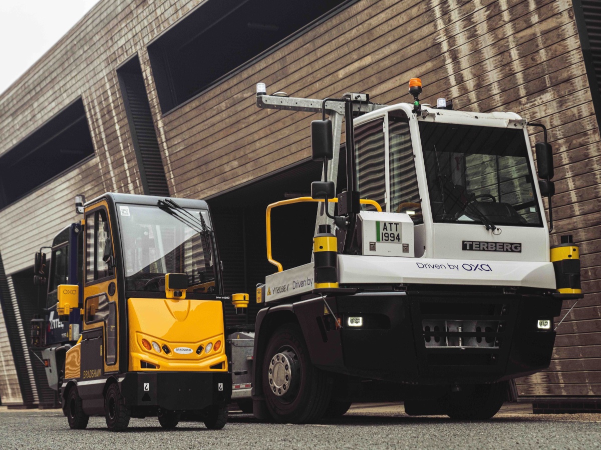 A large, white self-driving industrial vehicle and a small, yellow self-driving industrial vehicle, both branded for