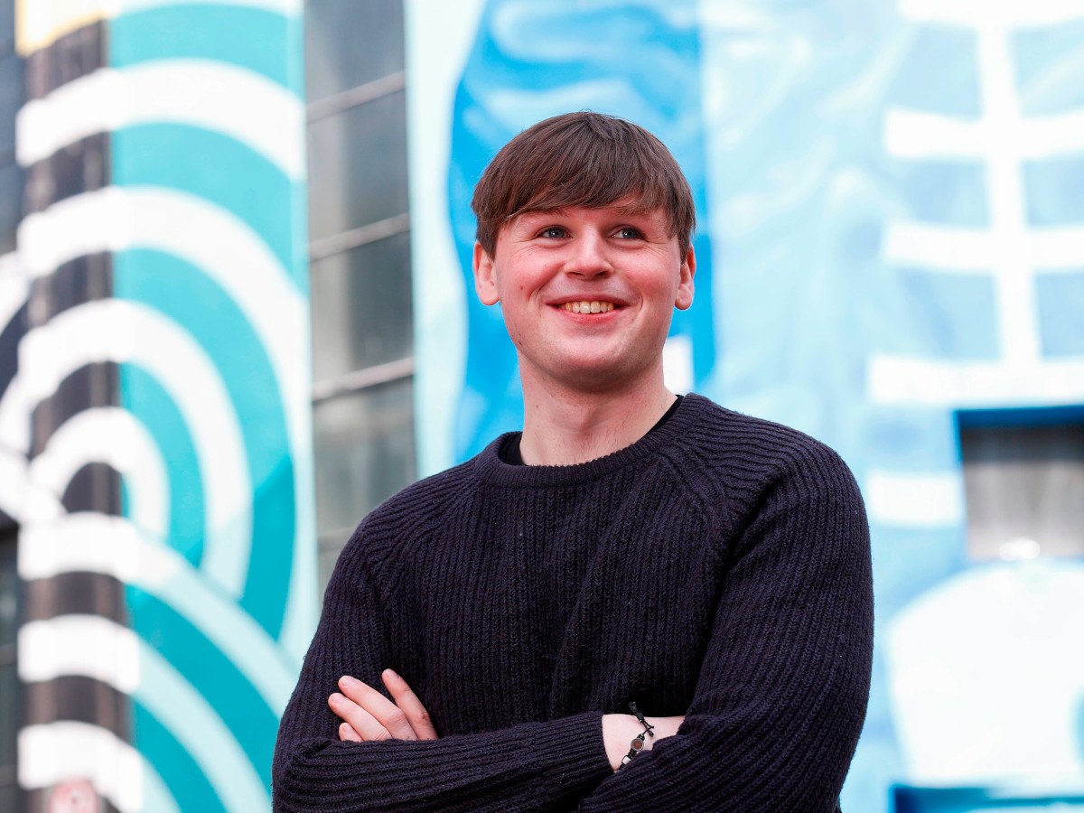 A man with short brown hair smiles with his arms folded wearing a black jumper.