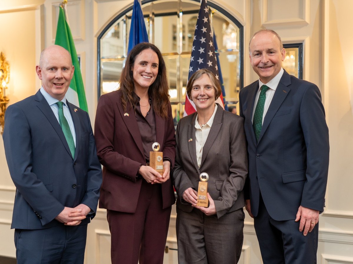 Two men and two women in business attire pose in front of flags.