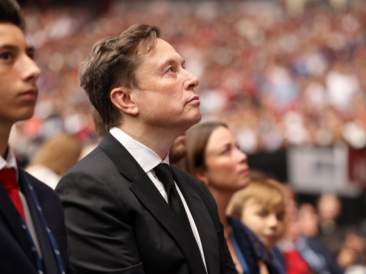 A man in a black suit and dark hair looking ahead. The photo has been taken to show his side profile. He is surrounded by others also looking in the same direction.