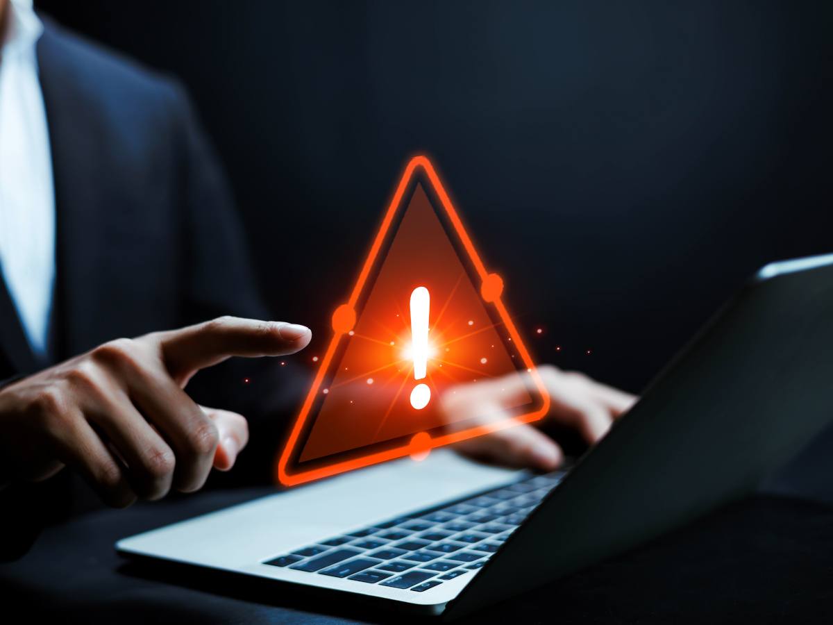 A hand reaches out to touch a red warning triangle hovering over a laptop signifying a cyber breach.