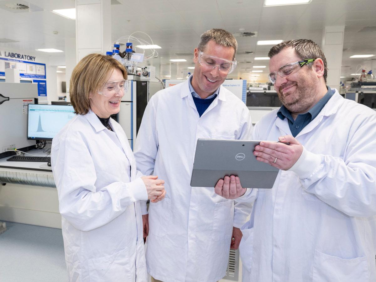 A woman and two men in lab coats, smiling and looking at a tablet in the hands of the man on the right.