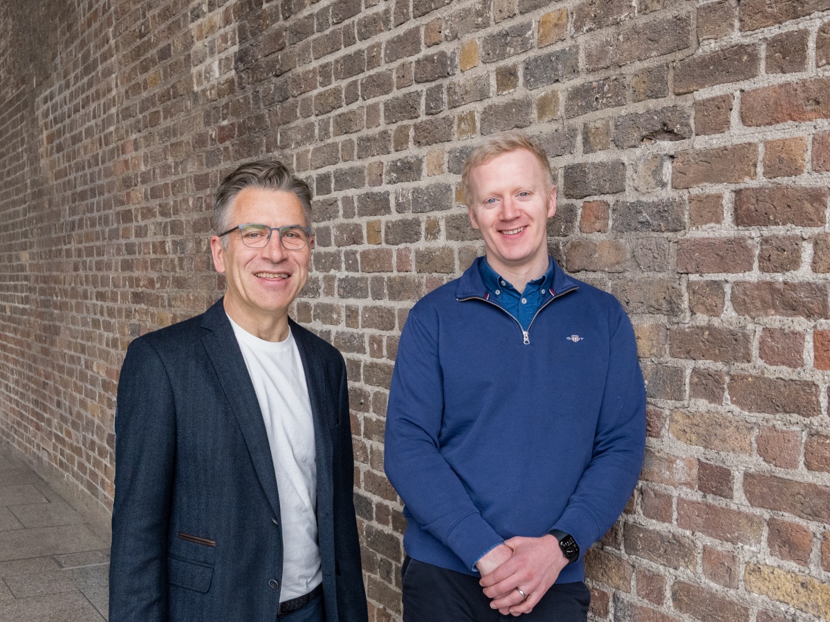 A white man in glasses and a navy blazer next to a red-haired white man in a blue jumper, leaning against a red-brick wall.