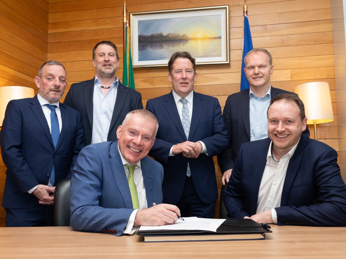 A group of six men in suits around a table. A man is sitting on a chair signing a documents on a table.
