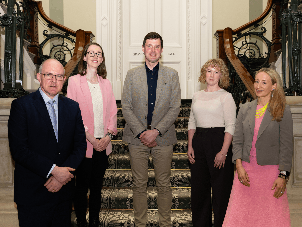 Two men and three women dressed professionally standing at the bottom of a fancy-looking staircase.