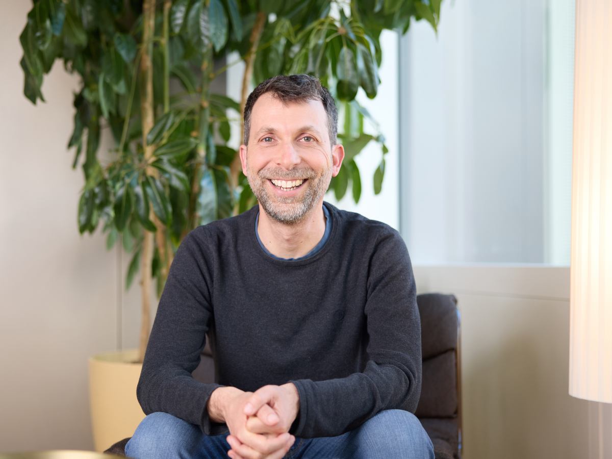 A white man in casual clothing poses, sitting and smiling, for the camera.