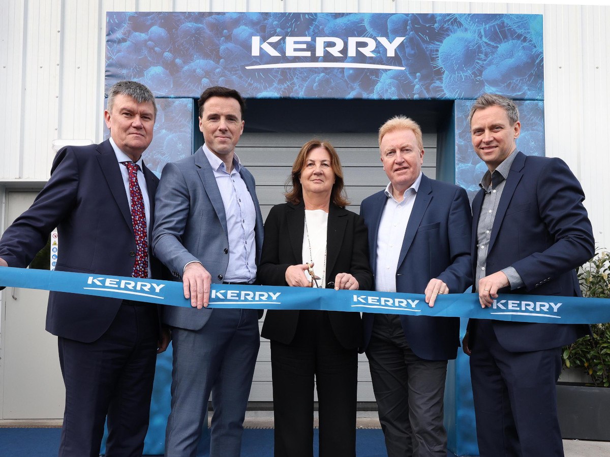 Five people in suits standing behind a ribbon, lightly holding it. The person in the centre is a woman with brown hair.