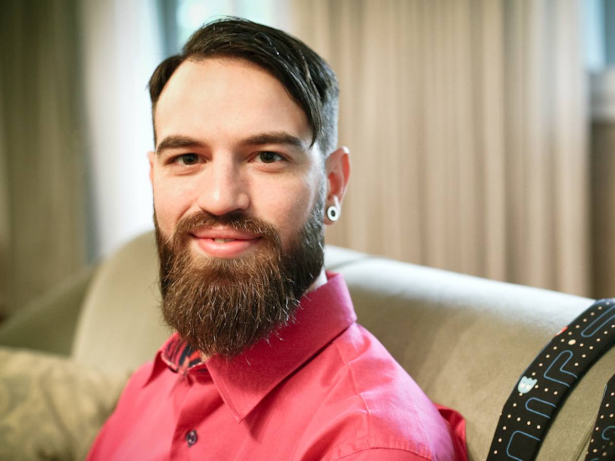 A bearded man in a pink shirt sits in a tan armchair