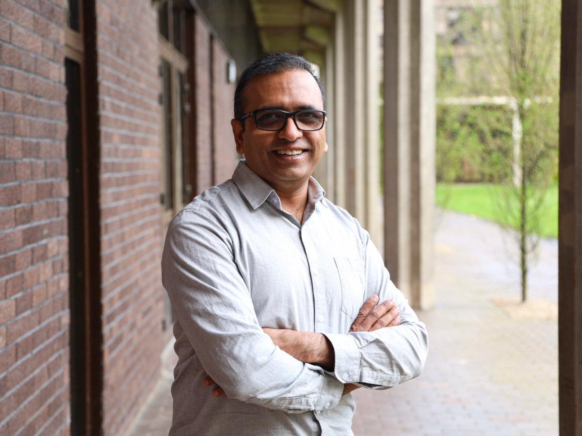 A man wearing glasses and a white linen shirt smiles with his arms folded while standing outside a red brick building.
