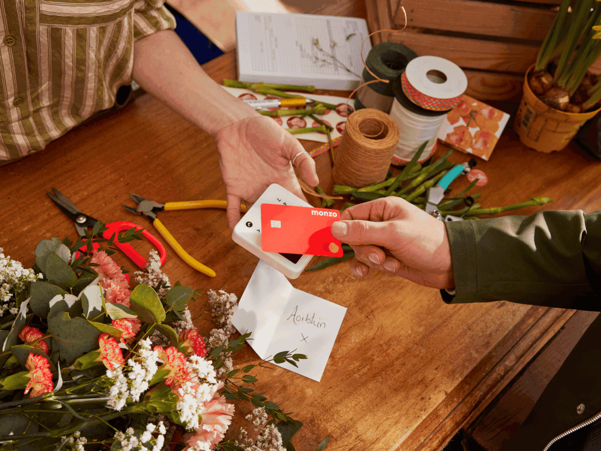 A arm is reaching out with a card reader and another arm is tapping their Monzo-branded card on the machine.