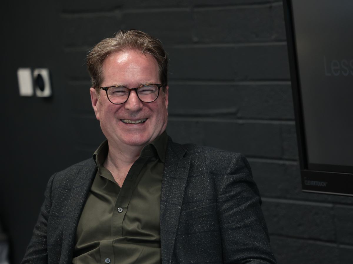 A man with glasses in a dark suit jacket, dark shirt, no tie laughing in conversation.