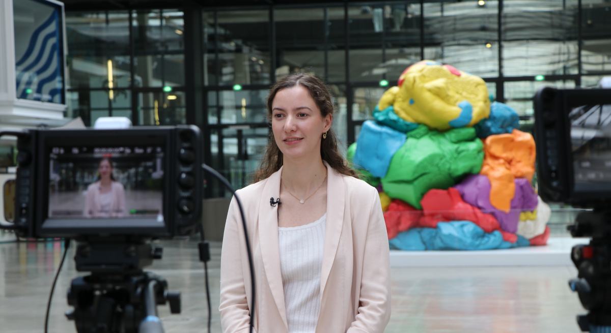A young white woman in a beige pants suit giving an interview in front of a recording camera.