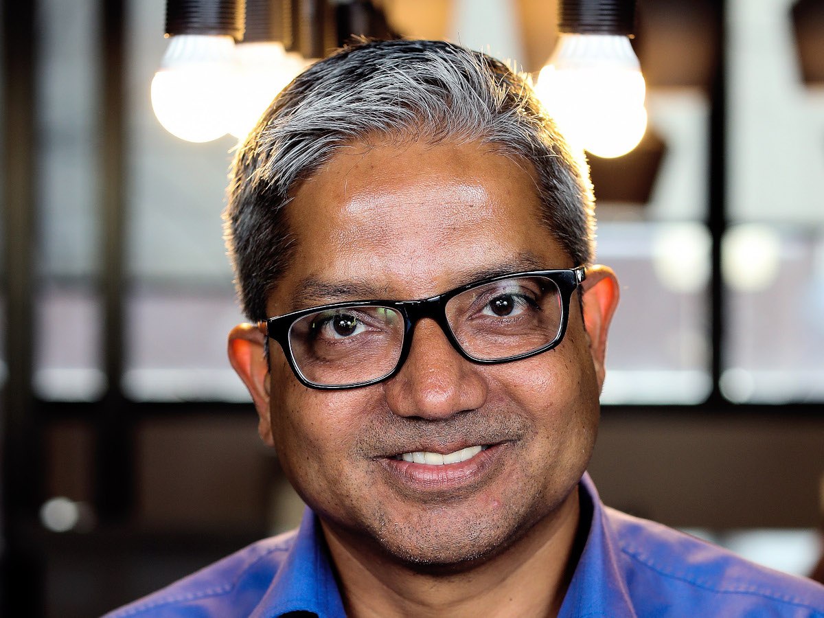 A man with dark, south-asian features, and grey hair with glasses smiling at the camera.