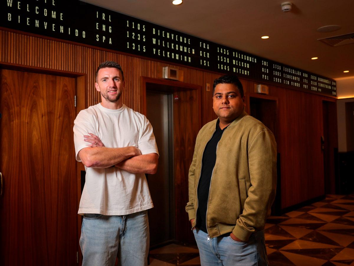 Two men, one wearing a white t-shirt with his arms folded and the other wearing a khaki jacket with his hands in his pockets, stand smiling in front of wood-panelled walls.