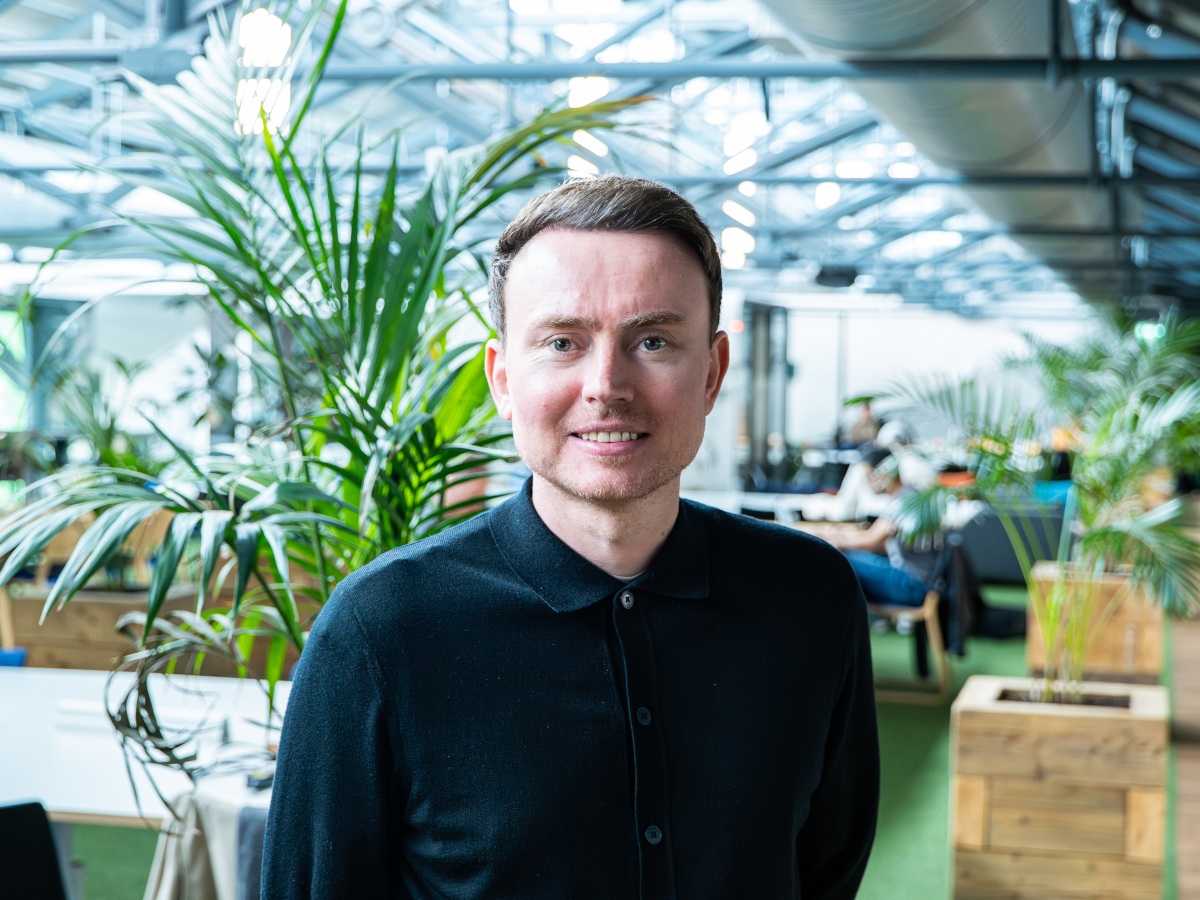 A young white man in a blue shirt, posing in an office with foliage.