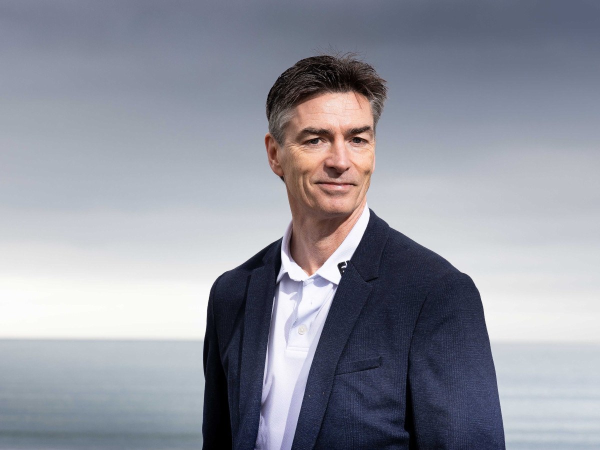 A middle-aged white man in a navy suit against a beach backdrop.