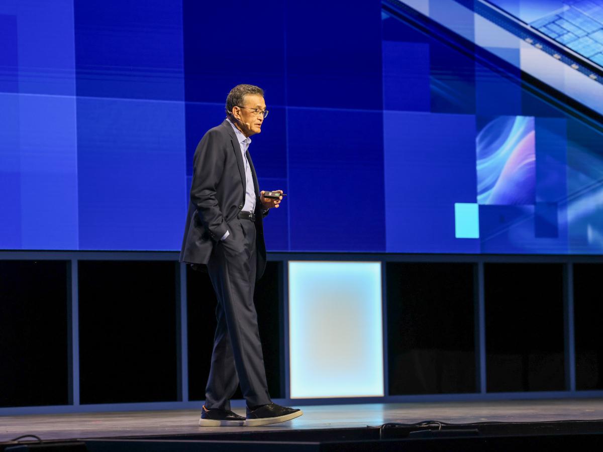 A man of Asian appearance walks on a large stage with blue background.