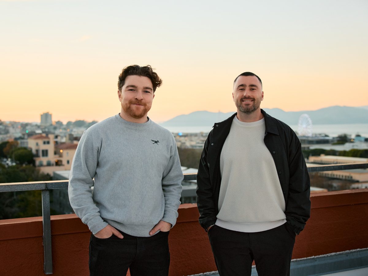 A man in a light blue sweater to the left and a man in a white T shirt and a black jacket over on top. They