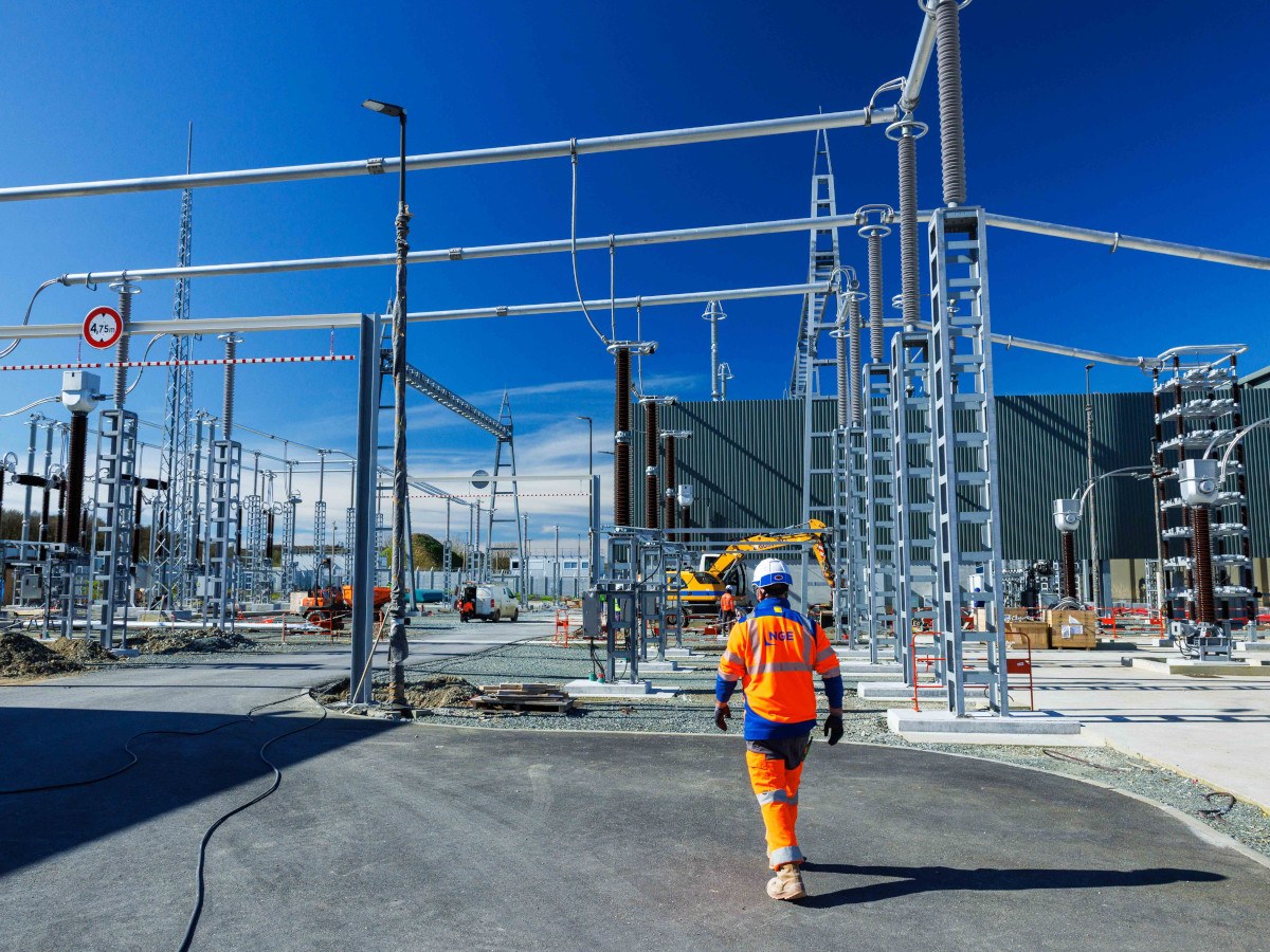 A man in orange construction gear walks among sea cable and energy infrastructure, with a blue sky in the background.