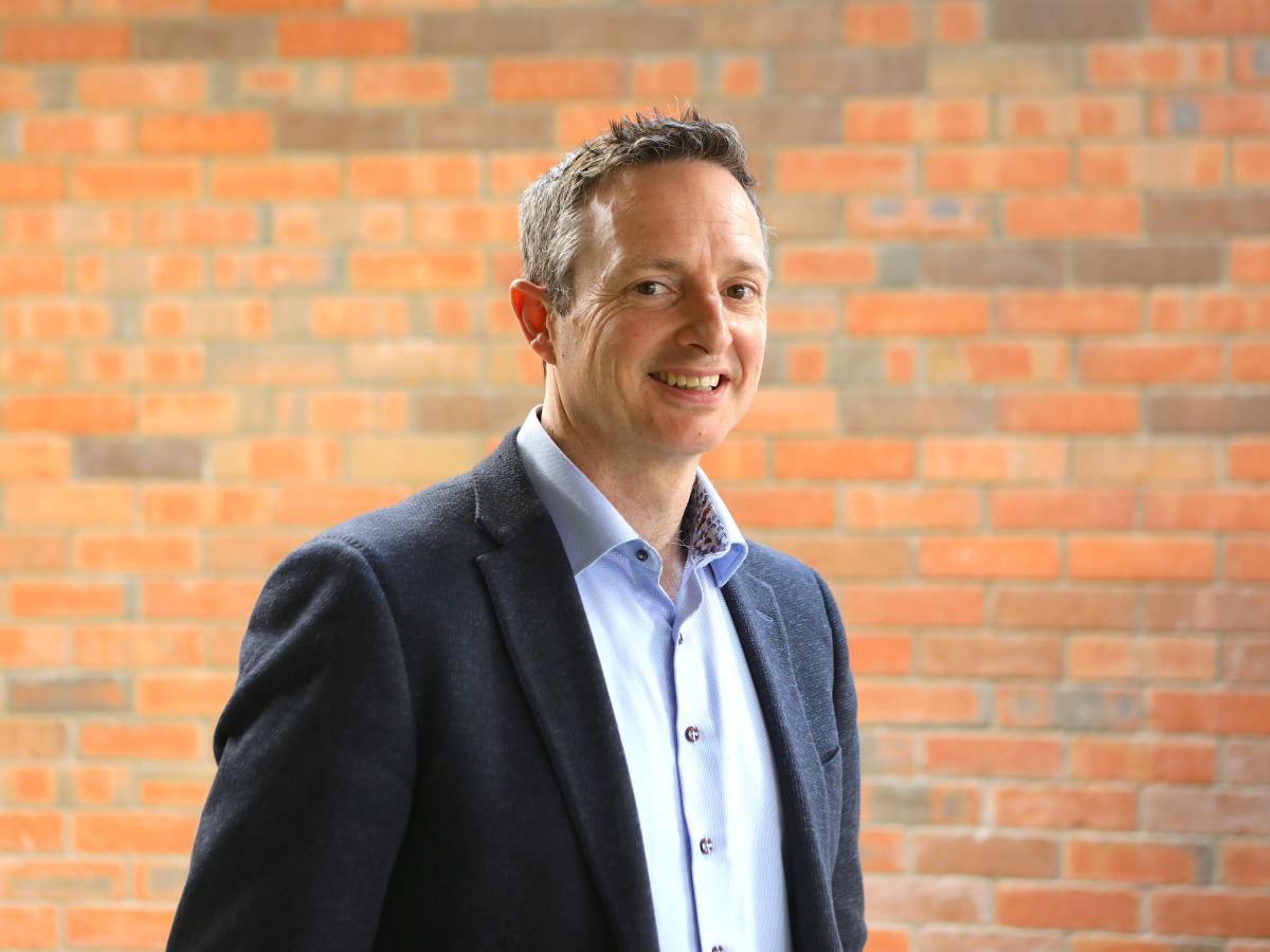 A man wearing a dark suit jacket over a light blue shirt with the collar open smiles in front of a red brick wall.