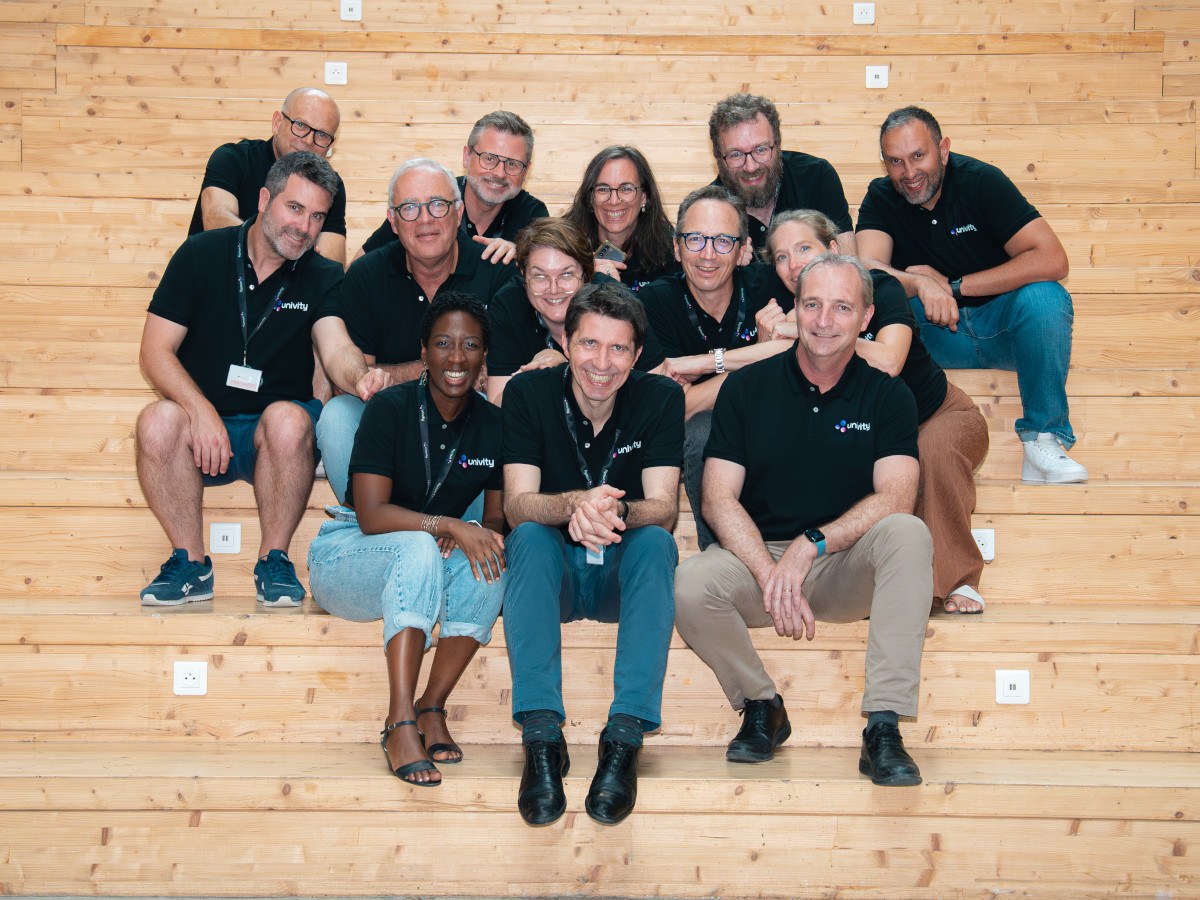 A group of 13 men and women sit on a set of wooden benches, all are wearing dark shirts with the Univity logo on them.