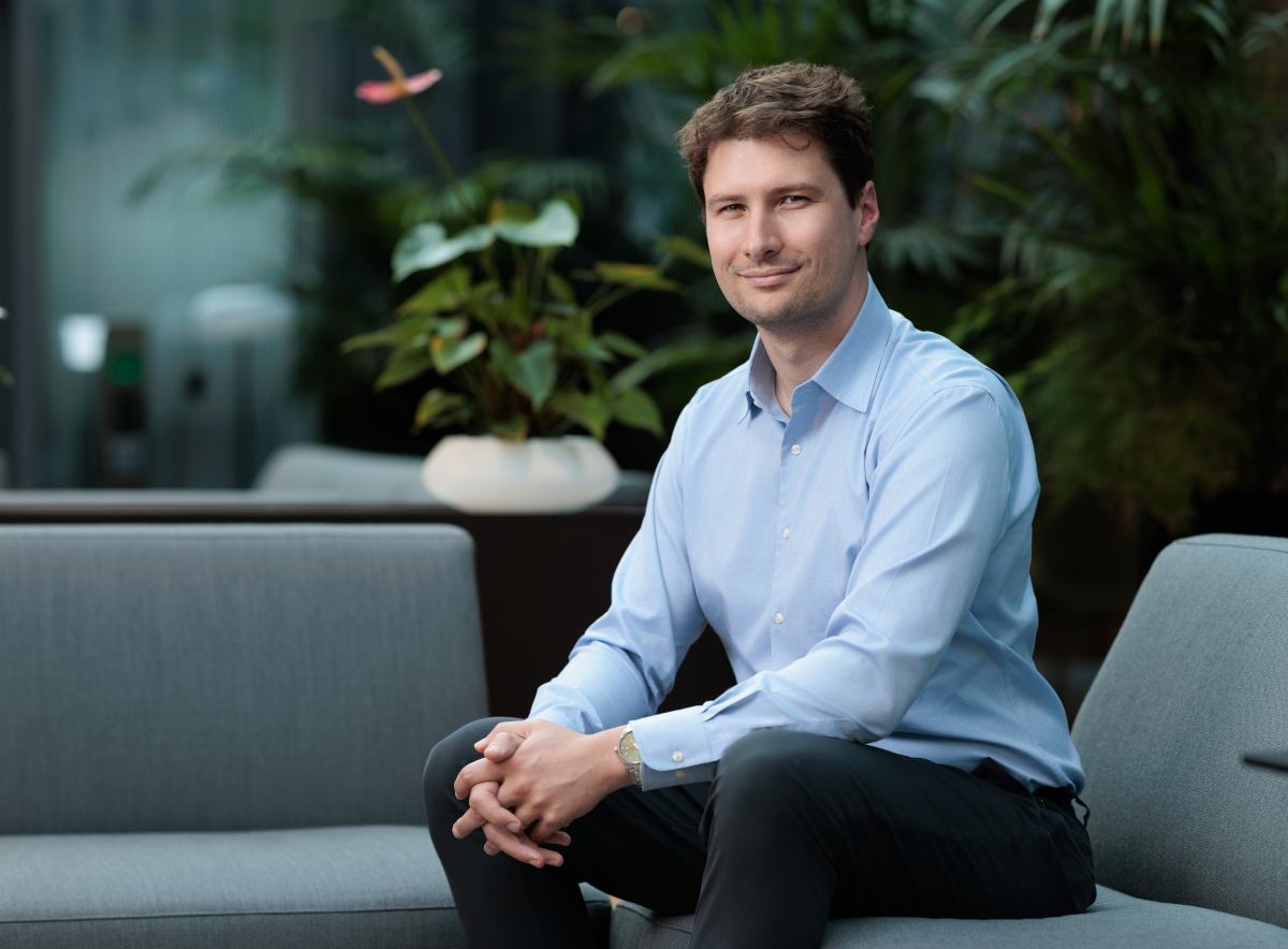 Caucasian man in blue shirt smiles to camera, seated on sofa.