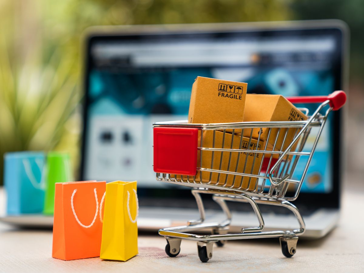 A mini shopping trolley filled with cardboard boxes with min shopping bags beside it. In the background is a laptop.