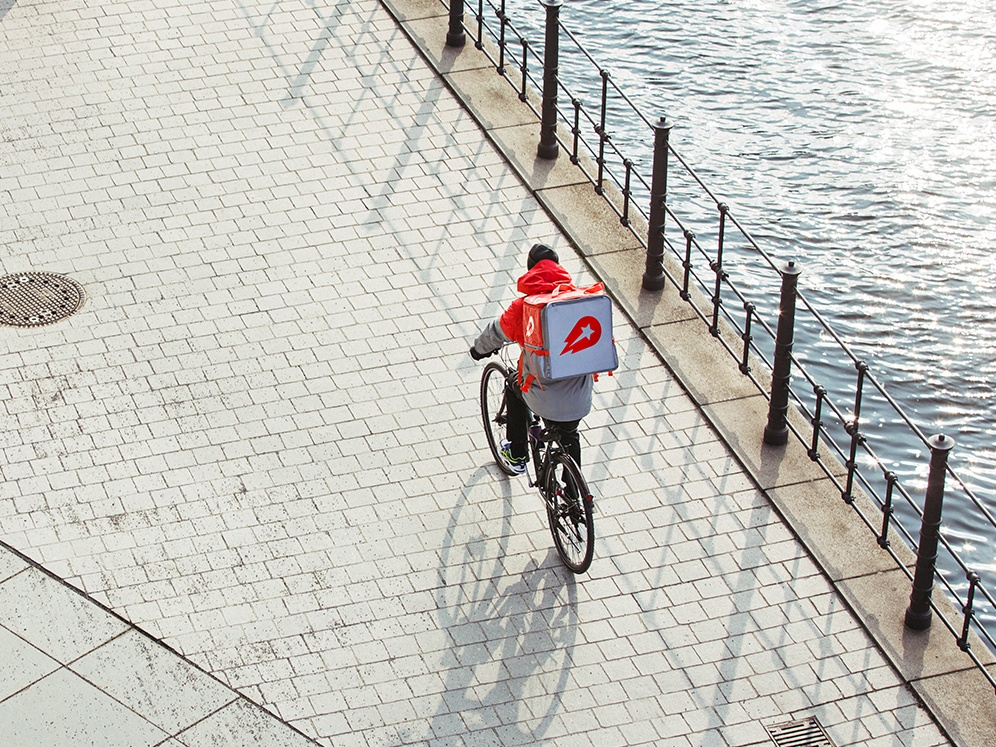 A person seen from afar wearing Delivery Hero uniform with a hot bag on a bicycle next to a body of water.