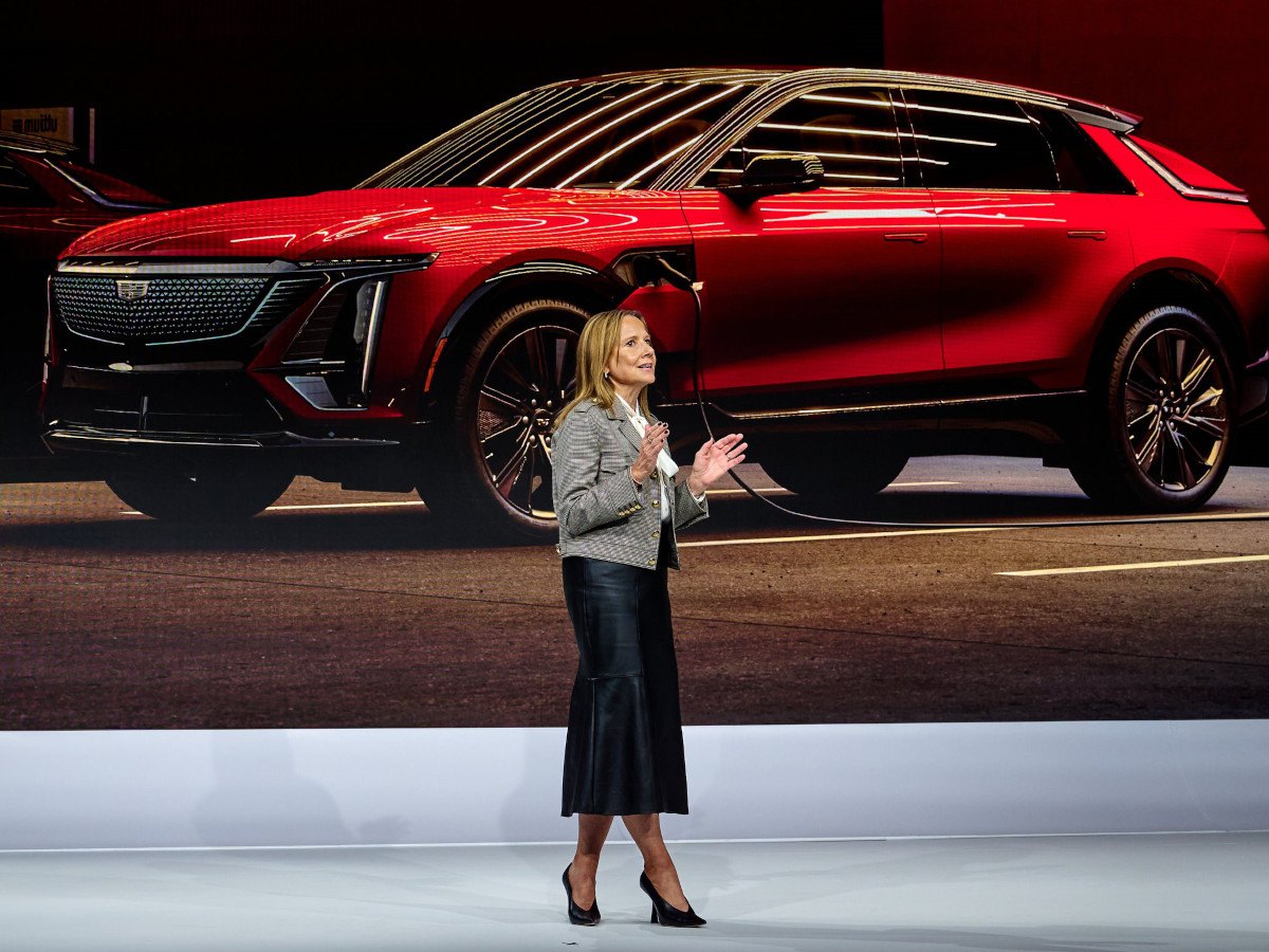 Mary Barra, the chair and CEO of GM standing, giving a presentation with a large red car in the background. Mary is wearing heeled shoes, a long skirt and a blazer.