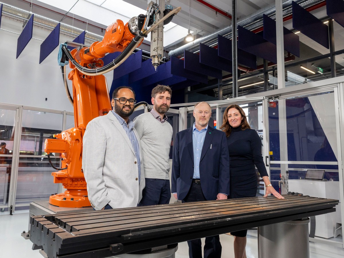 Four people standing together in front of what looks to be a work top with a large factory robot behind them.