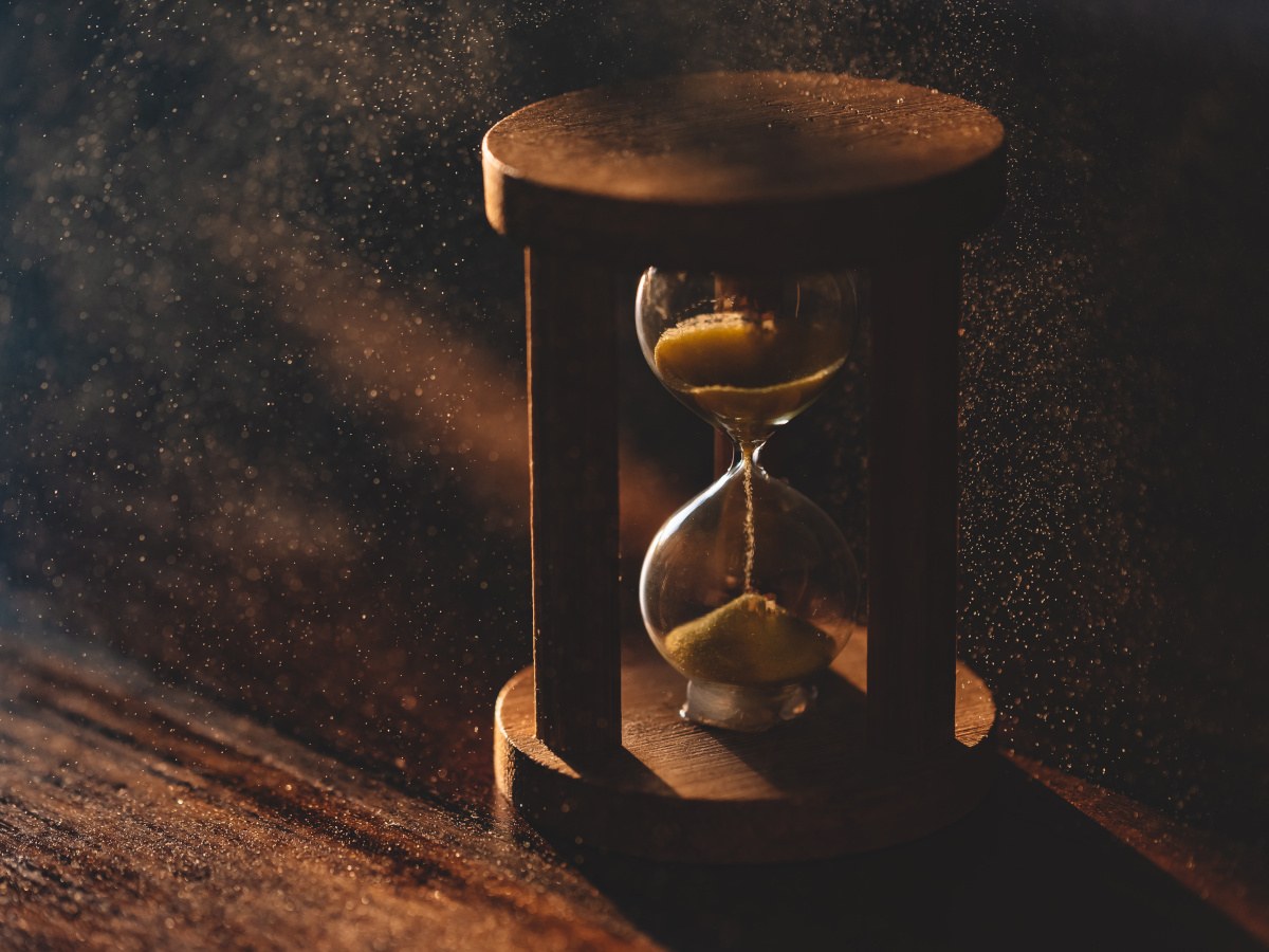 A picture of an old hourglass with sand drizzling down from the top part, sitting on a dusty wooden desk with minimal sunlight.
