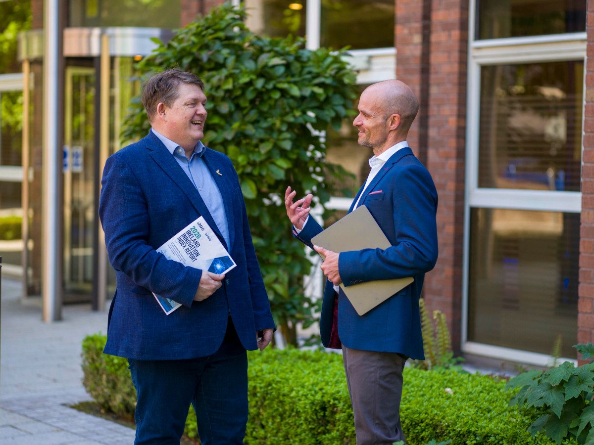 Two middle aged white men turn to each other and talk against a natural background.