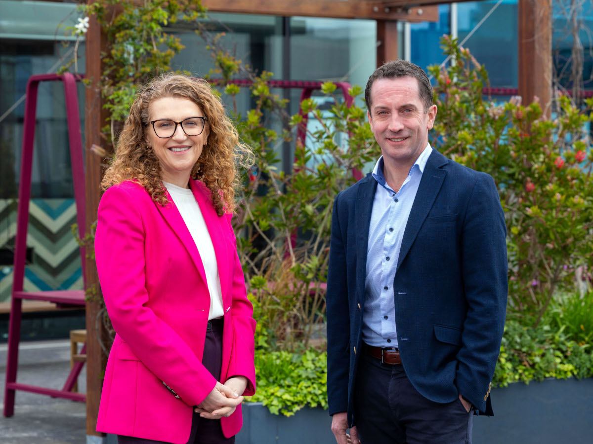 A woman in a pink blazer and black pants with a man in a blue suit posing in front of shrubbery.