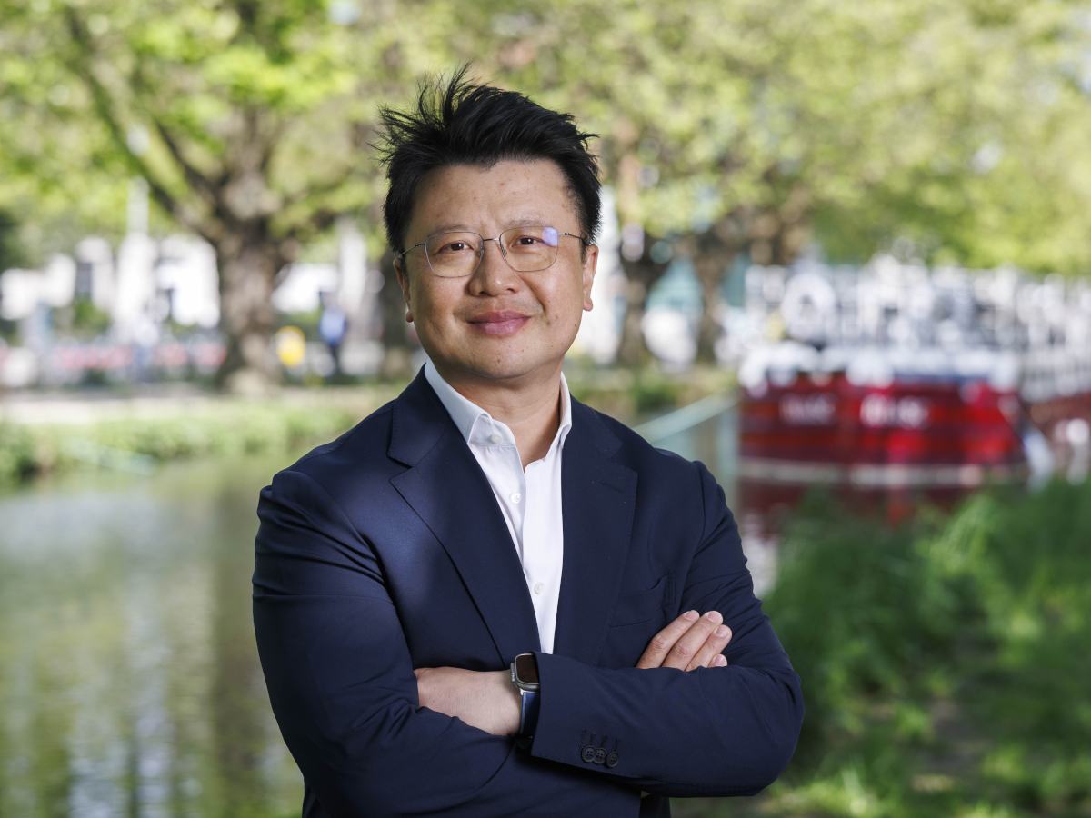 Man of Asian appearance with glasses and arms folded, leafy background